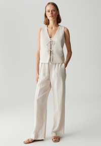 Lichtbeige, cropped vest met V-hals en strikdetail aan de voorkant, gecombineerd met losse, lichtbeige broeken. Reflecteert een gladde textuur en een minimalistisch ontwerp.