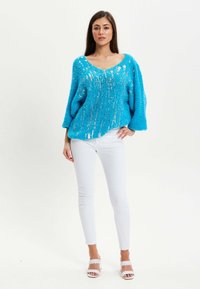 Bright blue oversized sweater with a V-neck, textured fabric, and metallic thread accents, paired with white skinny jeans and white sandals.