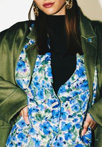 Gilet à imprimé floral aux tons bleus et verts, superposé sous un haut foncé et un manteau vert olive. Il présente de gros boutons et des coutures détaillées.
