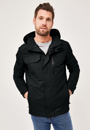 Schwarze Zip-Jacke mit Kapuze, die über Vorderasstaschen, einer strukturierten Oberfläche und einem leicht taillierten Design verfügt. Getragen über einem grauen Hemd.