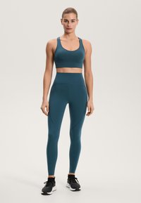 Türkizkék sportöltözet, amely formás sportmelltartót és íves varrású, magas derekú leggingset tartalmaz. A megjelenést fekete sportcipő teszi teljessé.