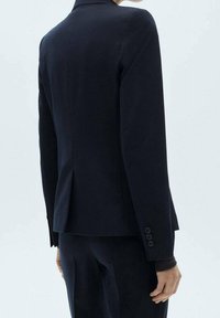 Blazer bleu marine cintré avec une silhouette ajustée, une fente arrière simple et des poignets à quatre boutons. Tissu lisse et design structuré.