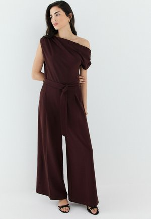 Frau steht in weit geschnittenem dunkelbraunem Jumpsuit mit einem Schulterärmel und gebundener Taille, trägt schwarze Riemchensandalen.