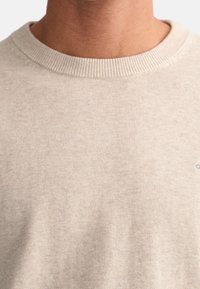 Beige gestrickter Pullover mit Rundhals, geripptem Kragen und subtiler Textur. Kleines Logodetail unten rechts.