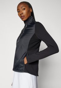 Černá sportovní bunda s vysokým límcem, zipem do poloviny a dlouhými rukávy; má jemnou, hladkou tkaninu a postranní kapsy.