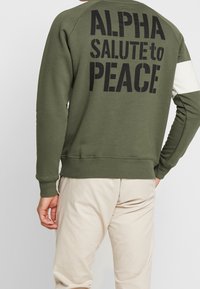 Felpa verde oliva con testo nero "ALPHA SALUTE to PEACE" sul retro. Presenta polsini e vita a coste, realizzata in morbido misto cotone.