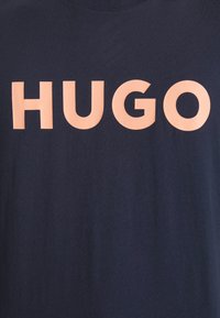 T-paita tummansinisestä puuvillasta, jossa on pyöreä pääntie, etuosassa suurilla vaaleanpunaisilla "HUGO"-kirjaimilla.