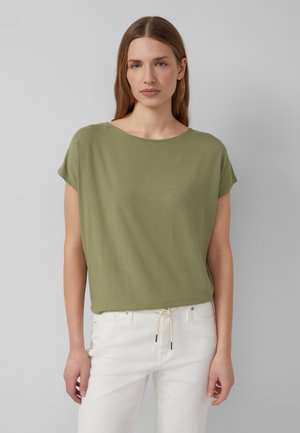 Vrouw met steil lichtbruin haar, gekleed in een losse, olijfgroene top met korte mouwen en witte broek met een taillekoord.