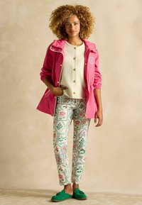 Jachetă impermeabilă roz cu glugă, purtată peste un pulover crem cu nasturi și pantaloni cu model floral. Mocasini verzi completează ținuta.