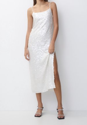 Kvinde iført en hvid midikjole med pailletter, tynde stropper og slids op til låret, kombineret med sorte hælede sandaler med stropper.