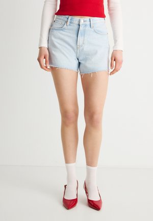Frau trägt hellblaue Jeans-Cutoff-Shorts, weiße Socken und rote spitze High Heels, steht vor einem schlichten weißen Hintergrund.