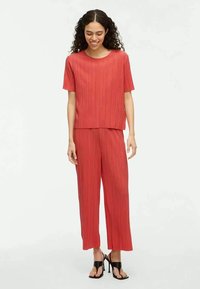 Red pleated two-piece outfit made of lightweight fabric, featuring a boxy top and wide-leg pants, complemented by strappy black heels.