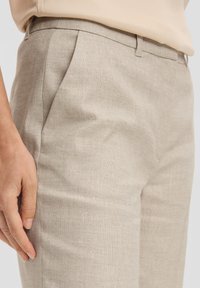 Beige linnenmix broek met een soepele textuur, voorzien van voren geplooide details en zijzakken, weergegeven met een hand die op de stof rust.