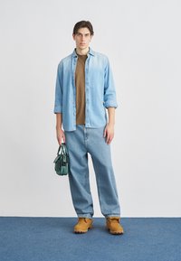 Νεαρό άτομο στέκεται κρατώντας πράσινη τσάντα χειρός, φορώντας ανοιχτό μπλε τζιν πουκάμισο πάνω από καφέ μπλουζάκι, φαρδιά μπλε τζιν παντελόνια και ταμπά εργασιακά μποτάκια πάνω σε μπλε χαλί.