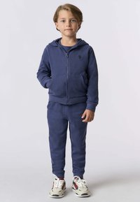 Un giovane ragazzo in piedi, che indossa una felpa con zip blu navy, pantaloni sportivi abbinati e sneaker bianche con accenti rossi e navy.