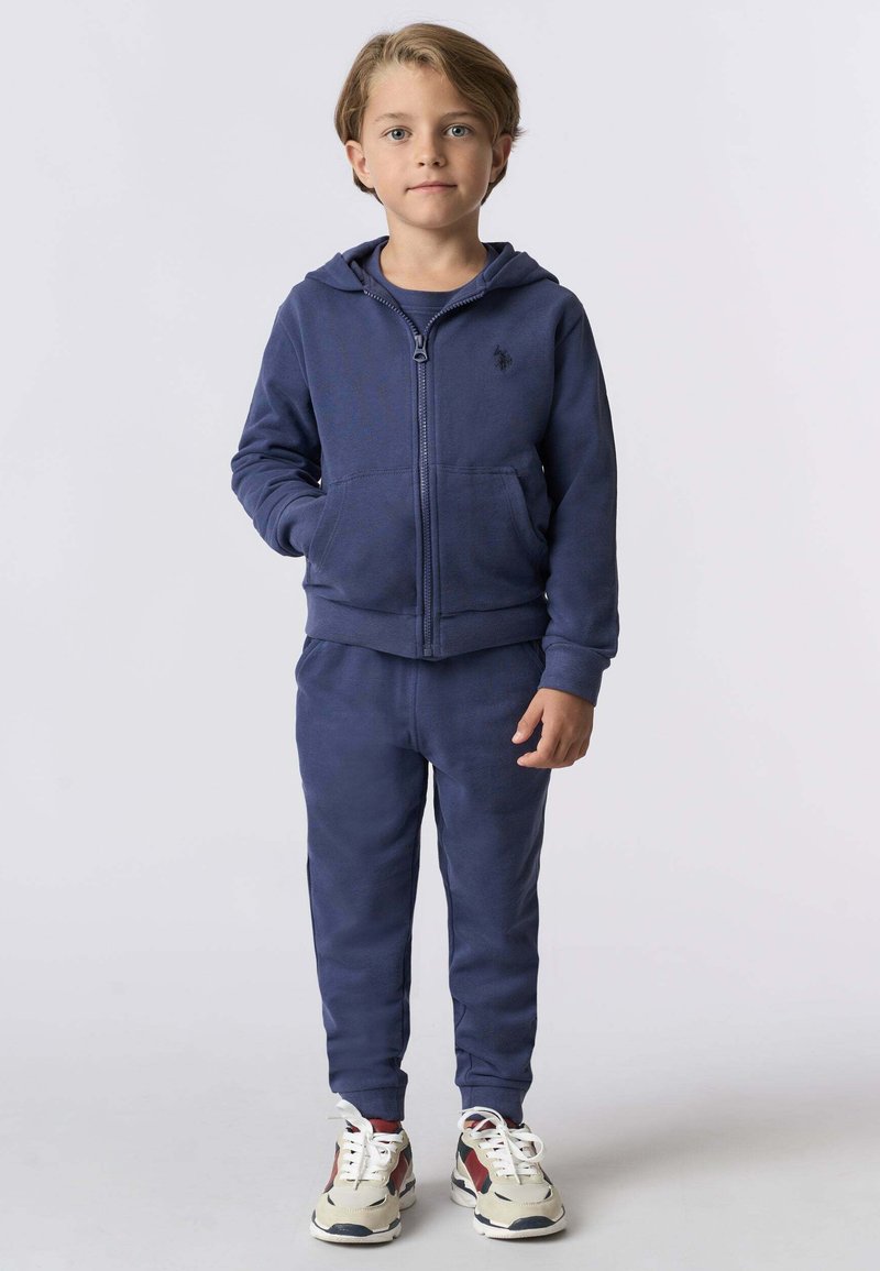 Un giovane ragazzo in piedi, che indossa una felpa con zip blu navy, pantaloni sportivi abbinati e sneaker bianche con accenti rossi e navy.