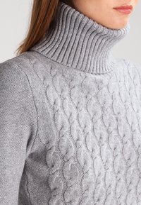 Grå stickad tröja med ribbad polokrage och flätstickad mönster på framsidan. Mjuk textur och åtsittande design.