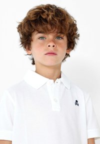 Polo blanco hecho de tela texturizada, con cuello y tapeta de tres botones. Destacado logo de calavera azul en el lado izquierdo del pecho.