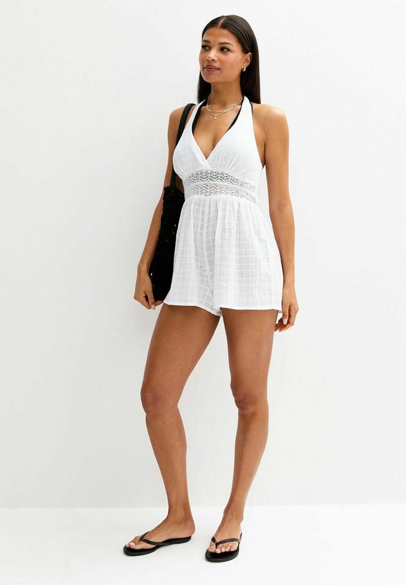 Witte halter-neklijf met een V-hals, textuurstof en kantaccent bij de taille, gecombineerd met zwarte flip-flops en een zwarte tote.