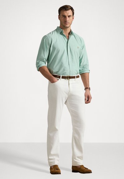 Gai zaļš krekls ar uzrullētām piedurknēm, baltas bikses un brūns jostas. Brūni loaferi noslēdz tērpu uz neitrāla fona.