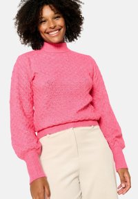 Maglione rosa lavorato a maglia con colletto alto, motivo a rombi e maniche a sbuffo. Indossato con pantaloni a vita alta, leggeri e a costine.