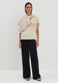 Top beige a maniche corte con dettaglio al collo annodato, abbinato a pantaloni neri a gamba larga. Texture a maglia liscia, design semplice, aspetto minimalista.