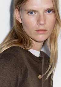 Hnědý cardigan s kulatým výstřihem a knoflíkovým detailem. Model má dlouhé světle hnědé vlasy a modré oči. Čisté, minimalistické pozadí.