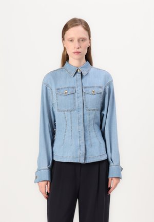 Chaqueta de denim azul claro con un diseño ajustado, dos bolsillos en el pecho con botones dorados y mangas largas con puños abotonados.