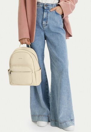 Osoba nosící světlé modré široké džíny a růžovou bundu, držící béžový kožený batoh za horní madlo.