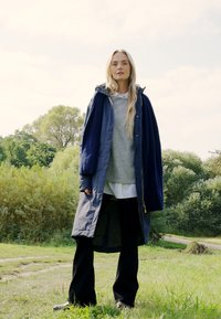 Lange marineblauwe jas over een grijze trui en wit shirt, gecombineerd met zwarte broek. De jas heeft een rits en is gemaakt van een zacht materiaal.