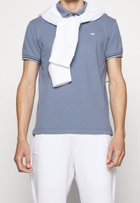 Camisa polo de algodón en azul claro con cuello y puños de canalé. Suéter blanco colgado sobre los hombros. Pantalones blancos ajustados.