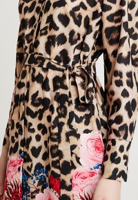 Robe à imprimé léopard en tissu soyeux avec manches longues, taille cintrée par une ceinture, et accents floraux rose et bleu sur l'ourlet. Détails de boutons visibles.