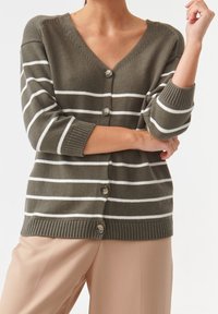 Osoba ubrana w oliwkowozielony kardigan w białe poziome paski z guzikami, w połączeniu z beżowymi spodniami, na jednolitym jasnym tle.