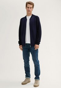 Granatowy sweter z dzianiny na zamek, z fakturą prążkowaną i kontrastowymi rękawami, zestawiony z jasnoniebieskimi dżinsami i beżowymi butami.