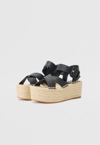 Sandali platform in pelle nera con cinturini incrociati, dettagli in corda di juta e cinturino alla caviglia con fibbia. La suola spessa aggiunge altezza e texture.