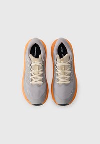 Baskets de course gris clair avec une tige en mesh texturé, lacets beiges et semelle en caoutchouc orange. Présente un logo sur le côté et la languette.