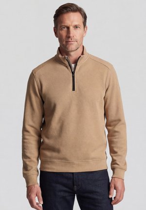 Man draagt een beige trui met een kwart ritssluiting en donkere spijkerbroek, staand tegen een effen lichtgrijze achtergrond.