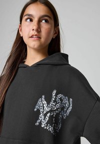 Čierna mikina s kapucňou a voľným strihom, na prednej strane so štruktúrovaným designom leopardieho vzoru v striebornej a modrej farbe.