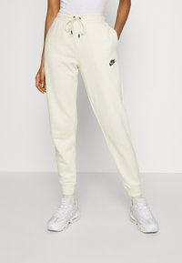 Joggers creme com um cós elástico, apresentando um logo preto da Nike. O tecido parece liso e suave, com punhos ajustados nos tornozelos.