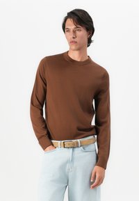 Homem com suéter castanho de mangas longas e jeans azul claro com cinto bege, de pé com uma mão no bolso contra um fundo branco.