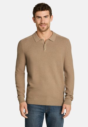 Homme portant un polo en tricot beige à manches longues et un jean bleu, debout les mains détendues le long du corps devant un fond uni.