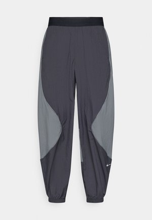 Pantalon coupe-vent Nike en gris foncé avec des panneaux latéraux gris clair. Taille élastique et bords aux chevilles. Tissu léger et texturé.