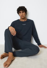 Maglietta a maniche lunghe blu scuro con logo sul petto, abbinata a pantaloni larghi coordinati. Tessuto morbido mostrato su uno sfondo chiaro.