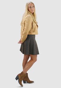 Beige leren cropped jack, grijze geplooide rok, bruine suède enkellaarzen met zwarte hakken, staande pose tegen een effen grijze achtergrond.