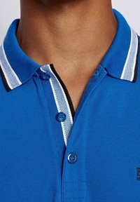 Blå polo-shirt med en tvåknappsknäppning, kontrasterande randig krage i vitt och svart, texturerad duk och broderad logodetalj.