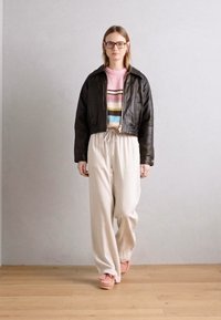 Jachetă scurtă din piele neagră, pulover tricotat multicolor cu dungi, pantaloni largi crem și pantofi roz deschis pe un podea din lemn.