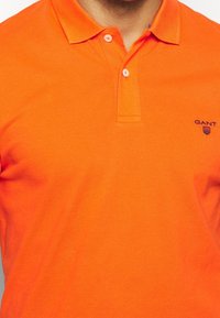 Jasnopomarańczowa koszulka polo z teksturowanej tkaniny, z klasycznym kołnierzykiem, dwoma guzikami i małym granatowym logo na lewej piersi.