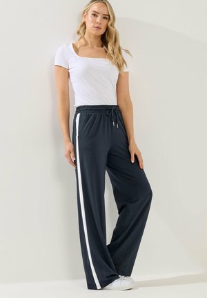 Vrouw staat tegen een muur, draagt een witte nauwsluitende top met korte mouwen, marineblauwe wijde broek met witte zijstrepen en witte sneakers.