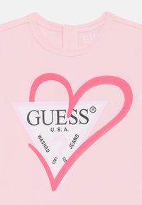 Rosa Baumwoll-T-Shirt mit einem großen Herz- und Dreieck-Logo in kontrastierendem Weiß und Rot, mit dem Text "GUESS U.S.A." und Produktdetails im Inneren.