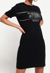 Robe t-shirt noire en coton, présentant un graphique "SUPERDRY" en blanc sur la poitrine. Manches courtes et coupe décontractée.
